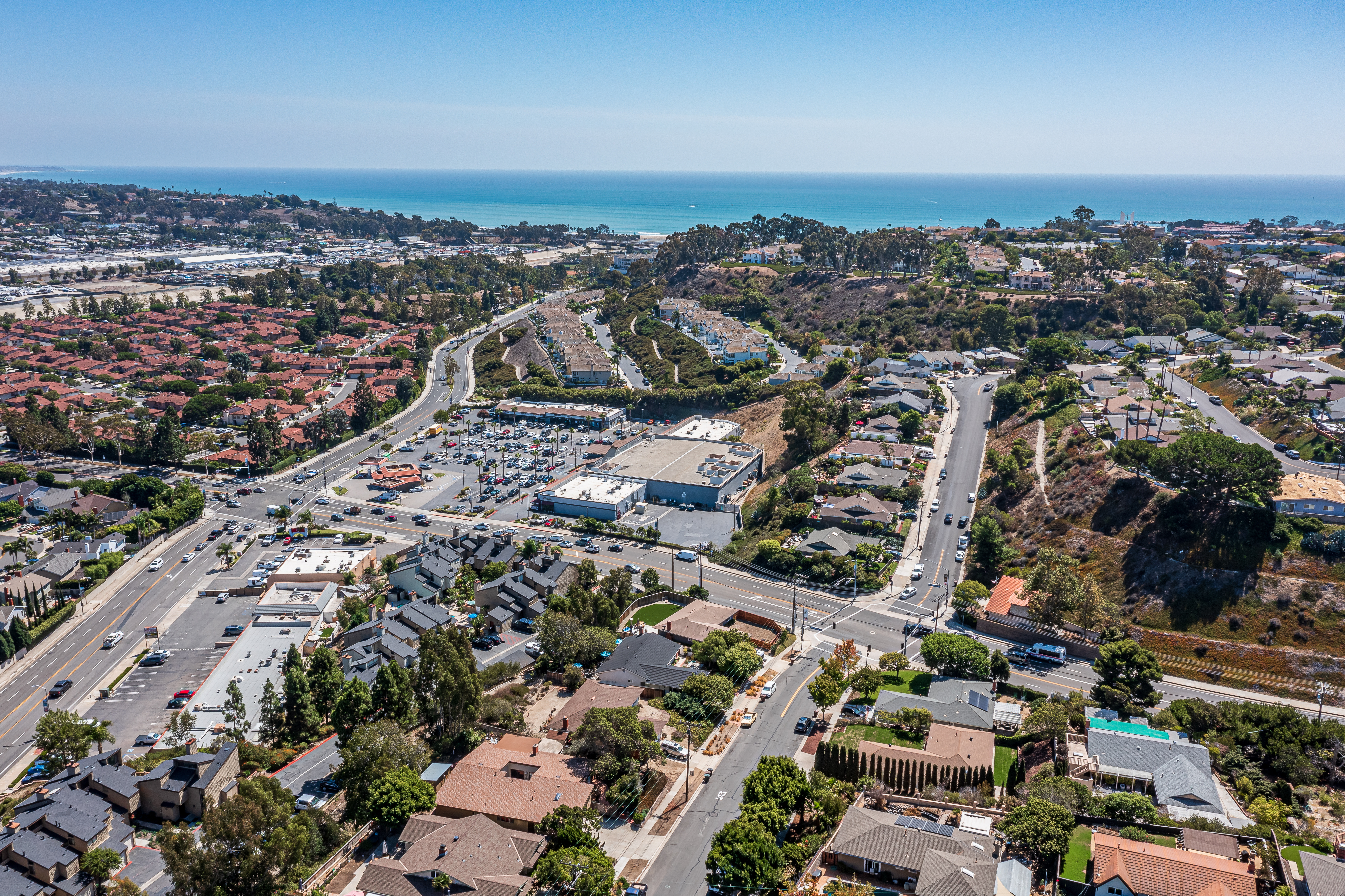 Aerial view of a coastal suburban area with residential homes, a shopping center, winding roads, and the ocean in the background under a clear blue sky.