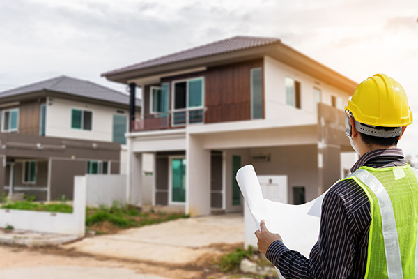 A construction worker in a yellow hard hat and reflective vest holds blueprints, facing a modern two-story house under a cloudy sky.